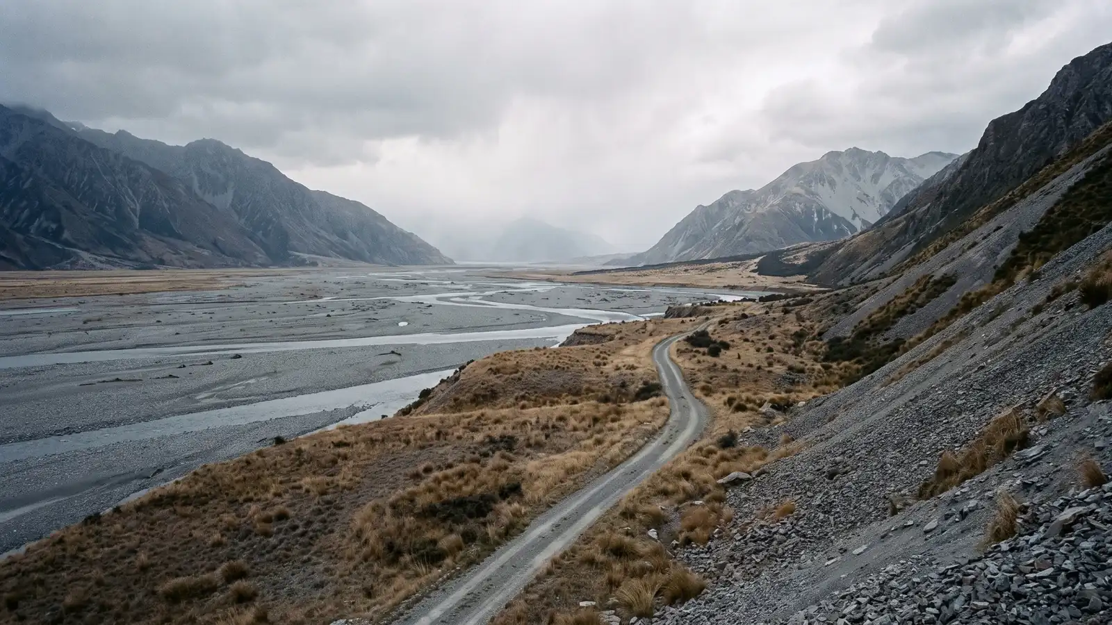 A braided gravel river valley in high country under flat overcast light with an unsealed exploration track along the terrace