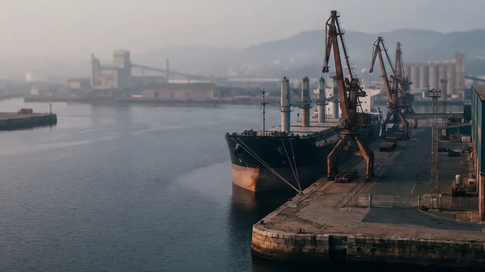 A bulk carrier moored at a coastal quay with dockside cranes and silos in morning haze