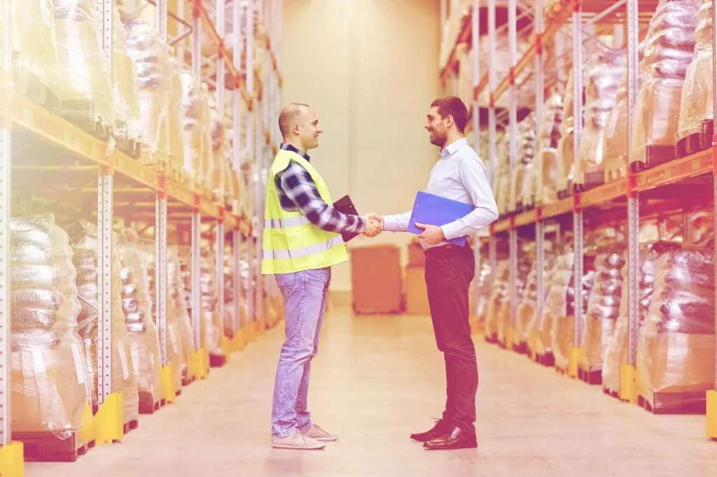 wholesale, logistic, people and export concept - manual worker and businessmen with clipboard shaking hands and making deal at warehouse