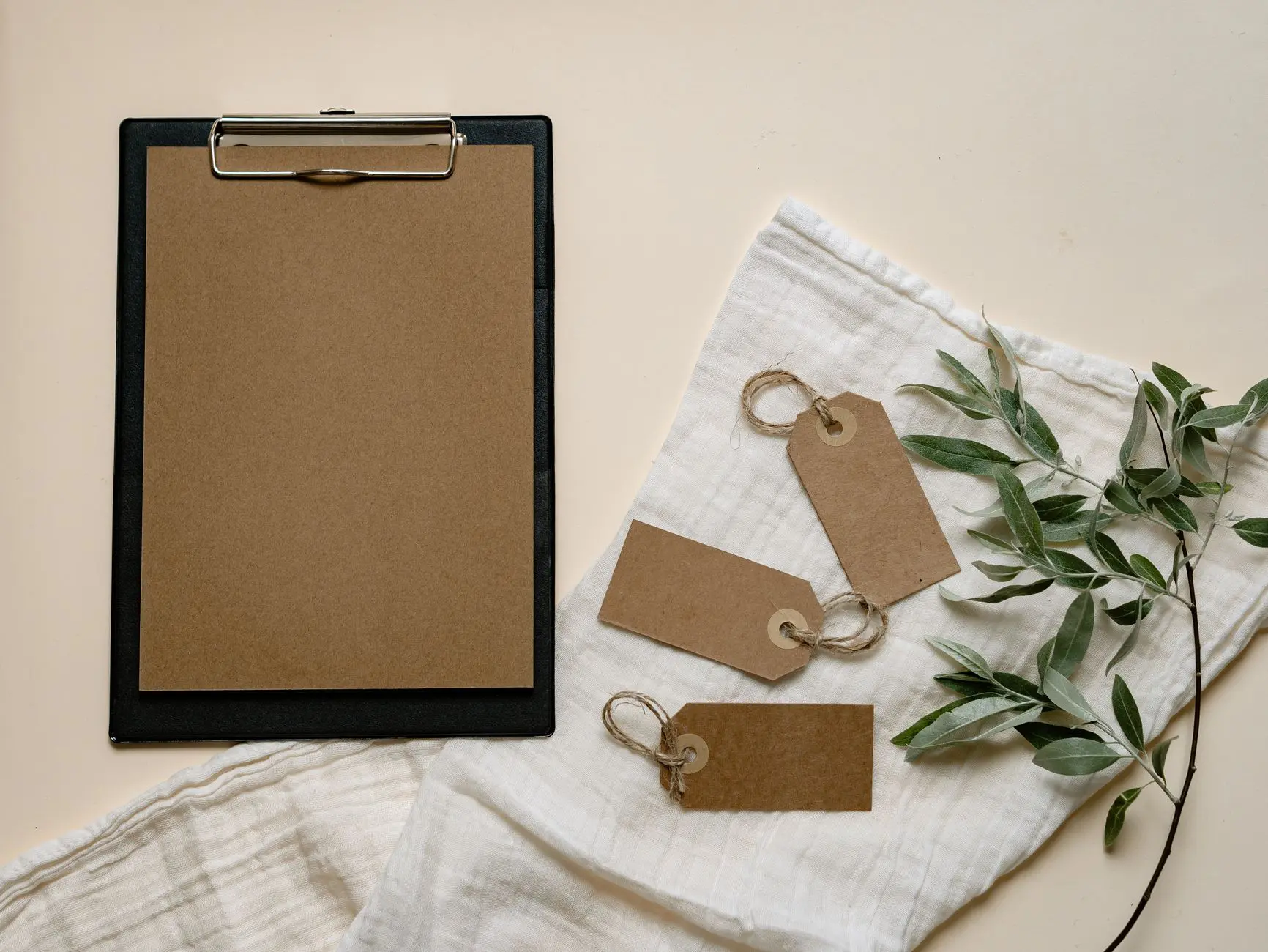 A minimalist flat lay of brown clipboard and tags with green leaves on white cloth.