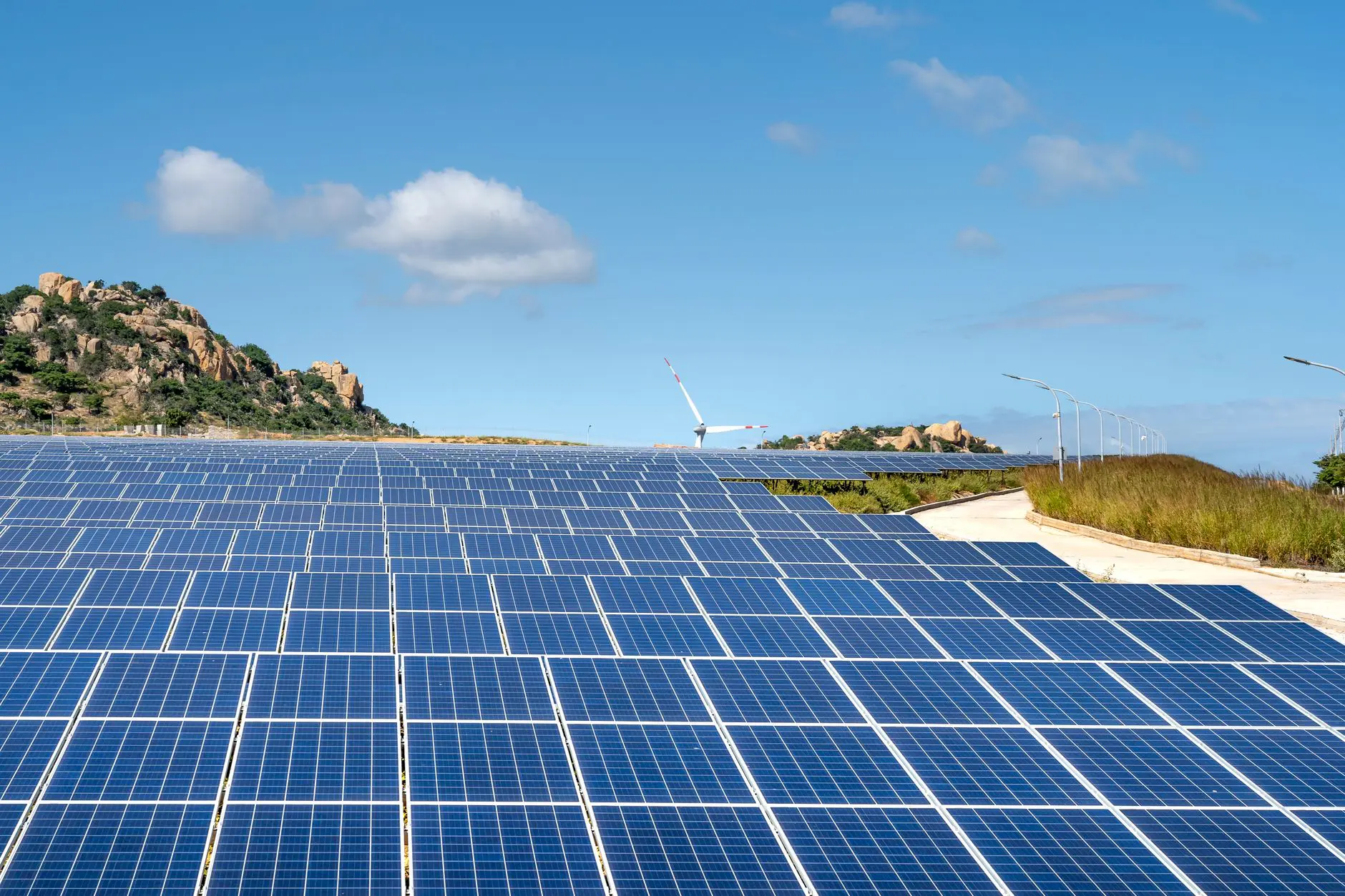 Expansive solar farm with rows of panels under clear blue sky and hills in the background.